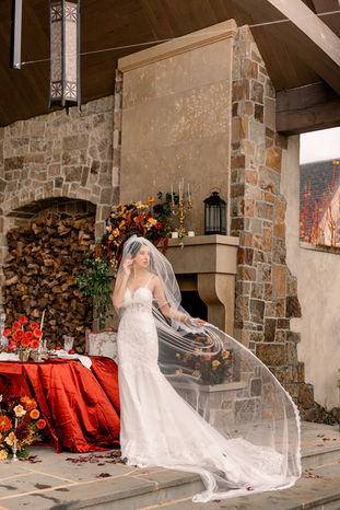 Bride beside an outdoor stone fireplace and firewood nook decorated with autumn blooms