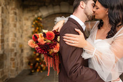 Groom kissing the bride's cheek as she holds a vivid autumn bouquet by a stone archway