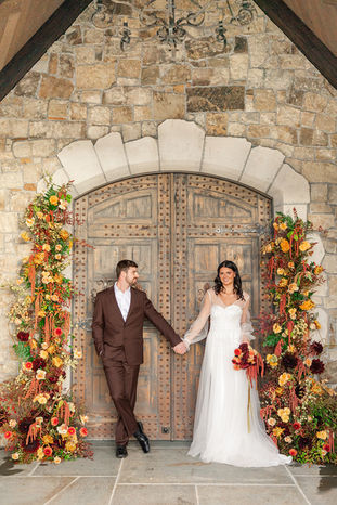 Bride and groom holding hands before the chateau's grand wooden door, flanked by floral garlands