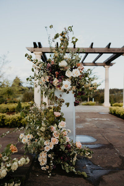 Lush ceremony floral arrangement of blush roses and greenery beside a stone pergola